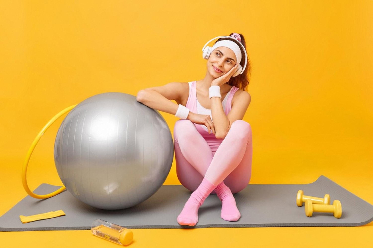Young woman in pink activewear resting on a yoga mat with fitness ball and dumbbells, illustrating post-procedure exercise routine